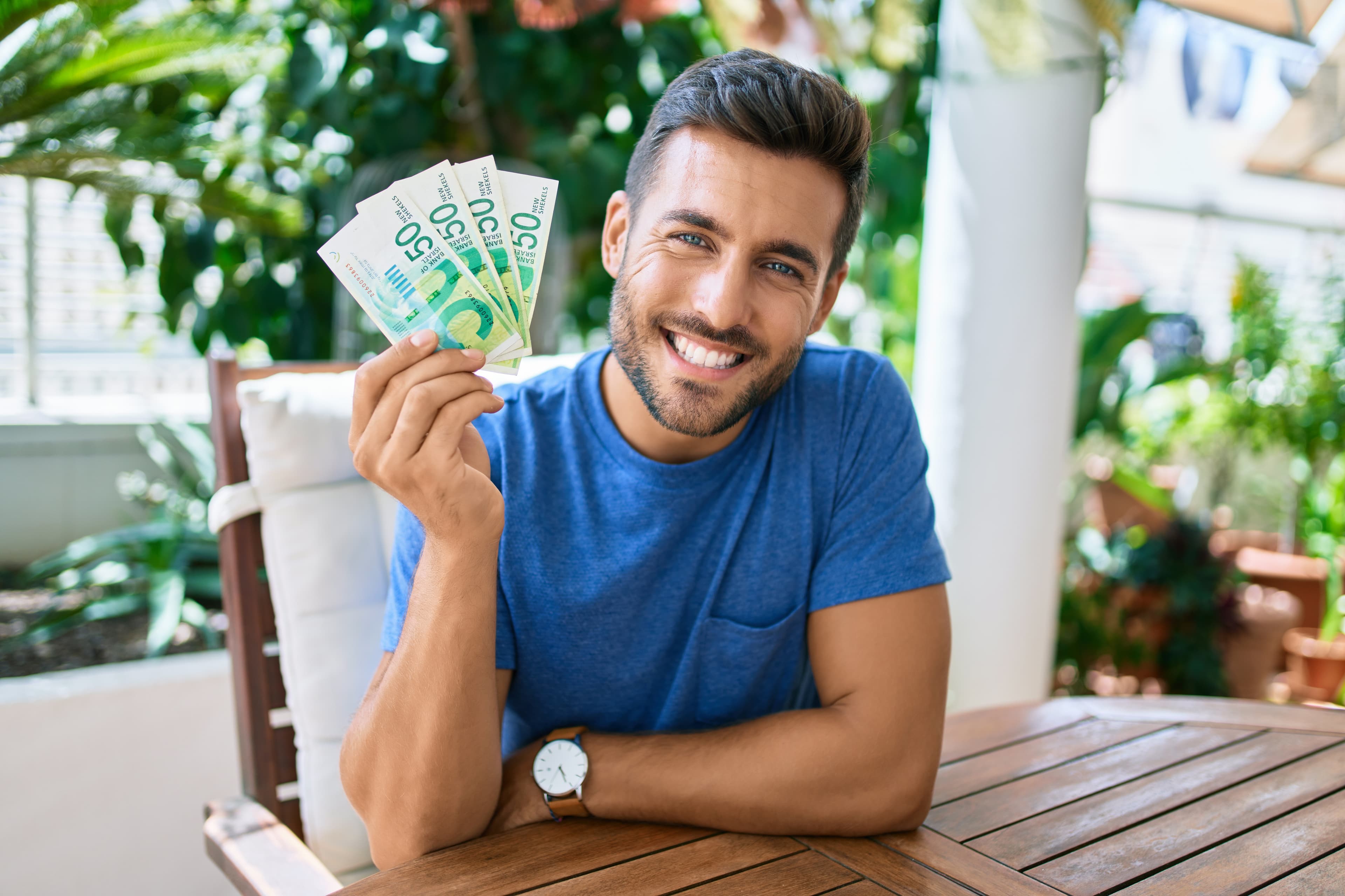 Smiling man holding shekels, sitting outdoors and looking pleased
