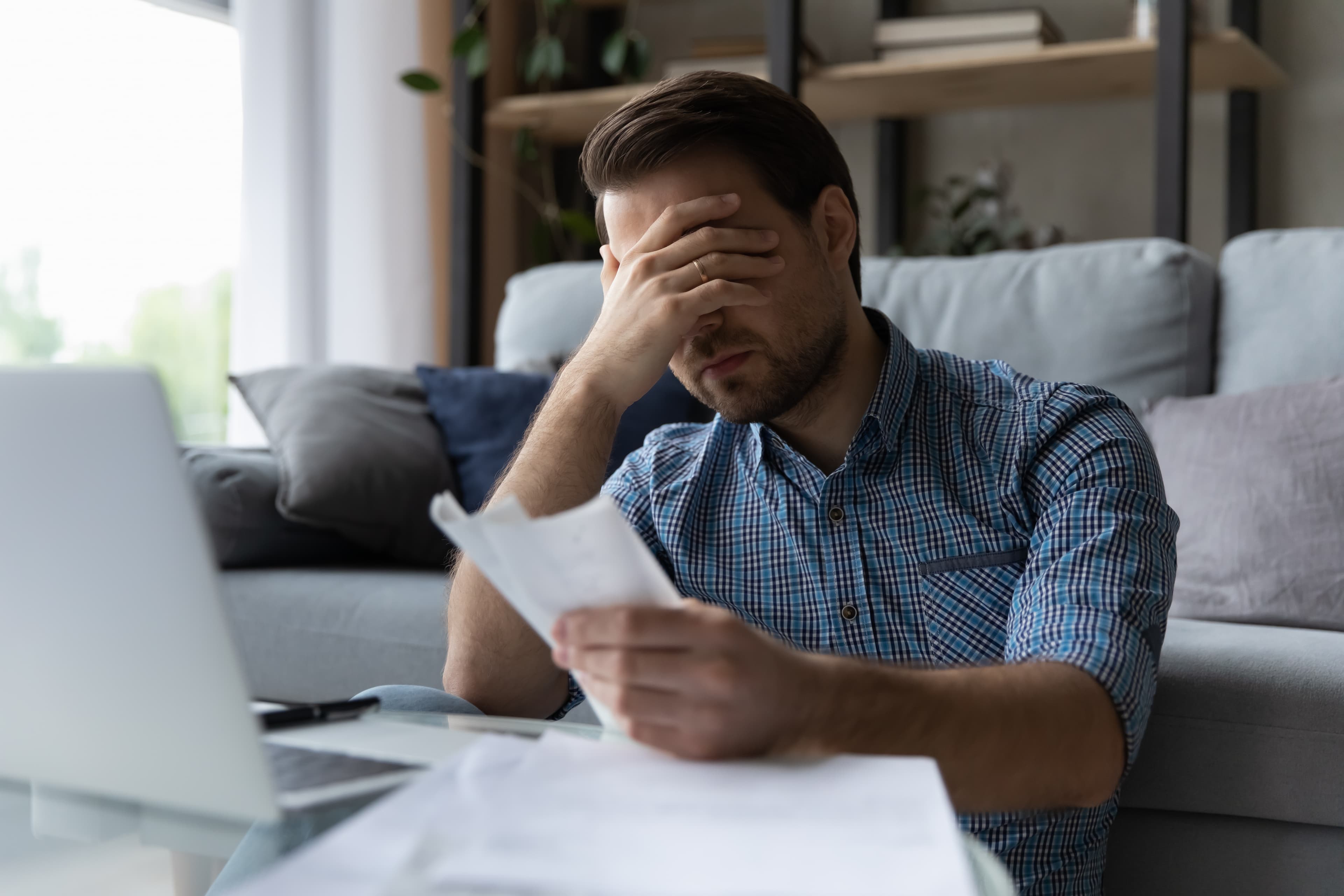 Frustrated man reviewing financial documents at home, holding his head in stress.