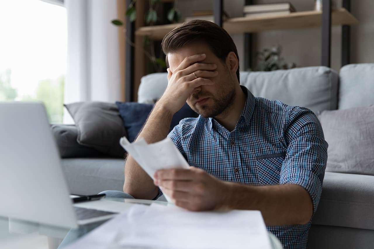 Frustrated man reviewing financial documents at home, holding his head in stress.
